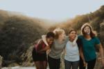 Four women with arms around each others shoulders laughing while walking in the dunes
