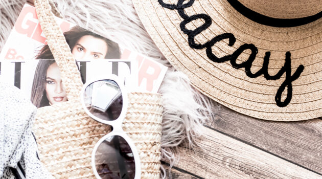 Beach bag with magazines sticking out with a pair of sunglasses on top and next to it a sunhat with Vacay embroided on the wide brim