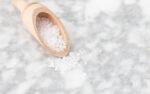 Close up of a wooden scoop with salt pouring out on a grey and white countertop