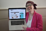 Bianca wearing a white shirt with a bright pink jacket, dark purple glasses and a patterned chemo hat holding her laptop