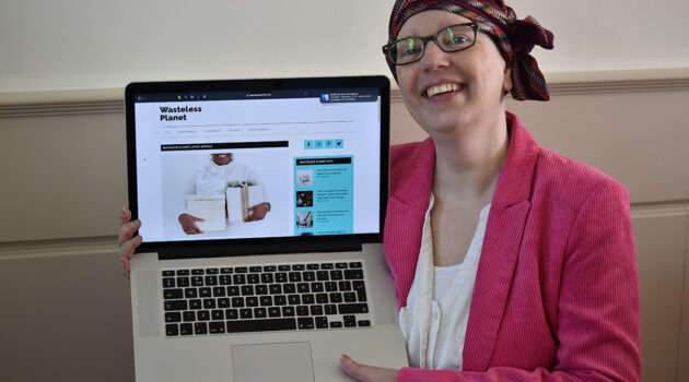 Bianca wearing a white shirt with a bright pink jacket, dark purple glasses and a patterned chemo hat holding her laptop